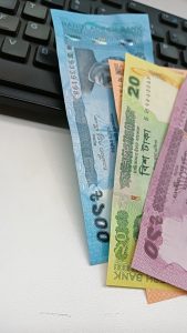 A close-up image of several banknotes, including a blue 500 Taka note, a green 20 Taka note, and a purple 50 Taka note, arranged on a white surface next to a black computer keyboard.
