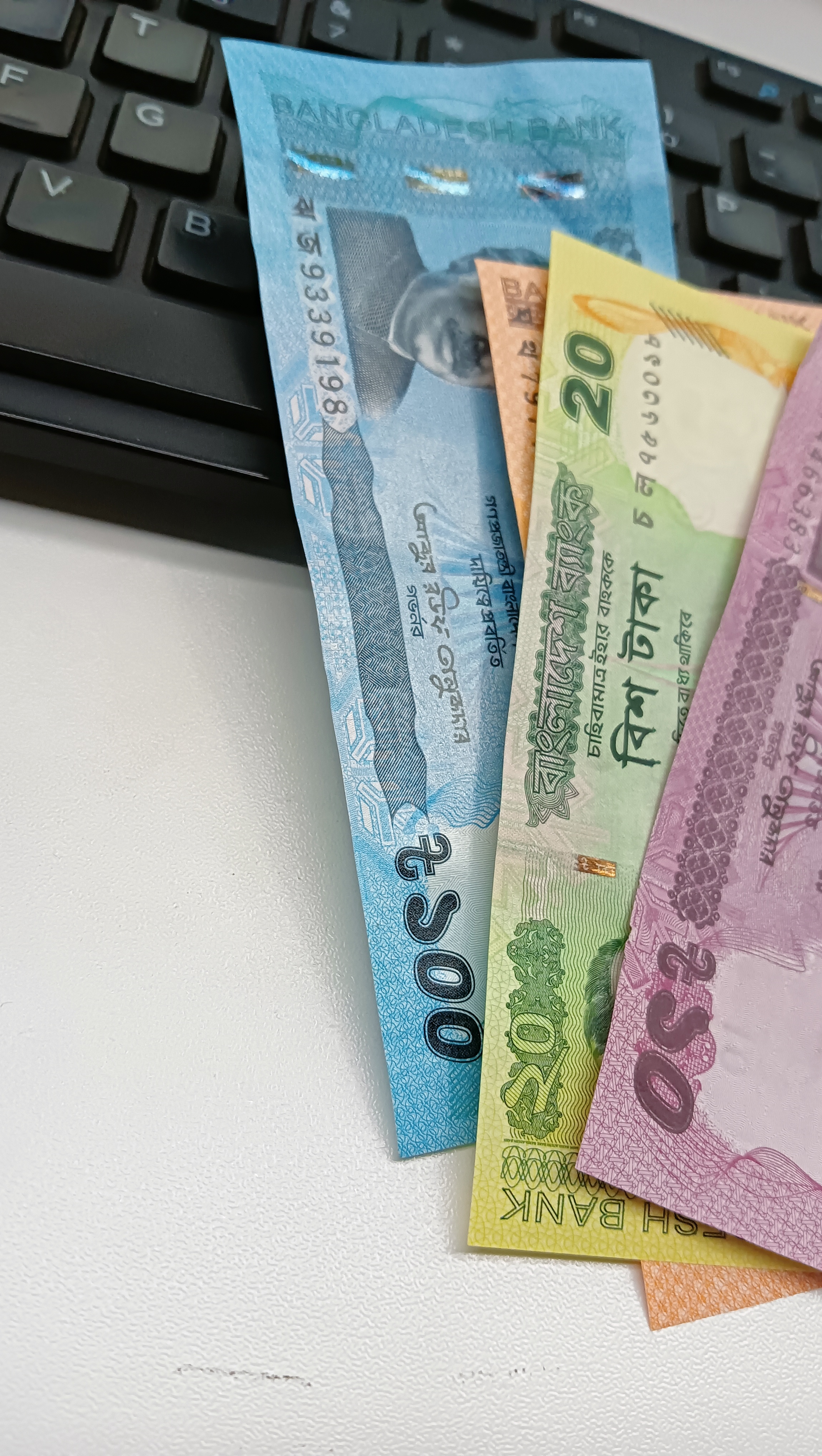 A close-up image of several banknotes, including a blue 500 Taka note, a green 20 Taka note, and a purple 50 Taka note, arranged on a white surface next to a black computer keyboard.