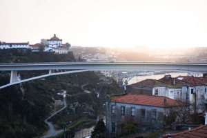 Sunset view of Porto, Portugal, showing red-roofed houses in the foreground, the Serra do Pilar Monastery on the hill, and modern bridges spanning the Douro River. 