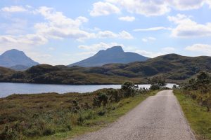 The Wee Mad Road winds past a sunlit loch, with hazy mountains towering on the way to Lochinver.