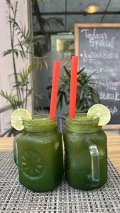 Two mason jar glasses filled with a green beverage, each adorned with a slice of lime.The background features greenery and a chalkboard displaying the day&#039;s specials.