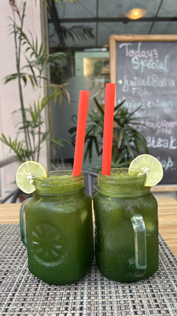 Two mason jar glasses filled with a green beverage, each adorned with a slice of lime.The background features greenery and a chalkboard displaying the day’s specials.