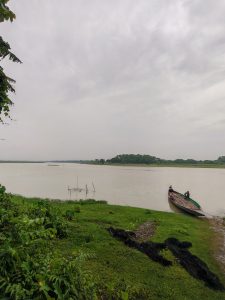 A serene riverside scene featuring a calm, muddy river under a cloudy sky. On the left, lush green grass and shrubs border the water, with a fishing net laid out on the ground. In the foreground, a wooden boat is docked by the shore, and a person is seen working on the boat, surrounded by gentle hills and trees in the background.