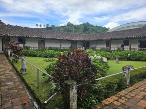 Colonial courtyard with hydrangea and tropical plant gardens, surrounded by tile-roofed corridors and white walls, framed by green mountains in the background under a partly cloudy sky.
Patio colonial con jardines de hortensias y plantas tropicales, rodeado de corredores con techo de tejas y paredes blancas, enmarcado por montañas verdes al fondo bajo un cielo parcialmente nublado.
