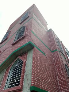 A corner view of a building featuring a red brick exterior with green trim. The structure has several arched windows with metal bars, showcasing a mix of modern and traditional architectural elements.