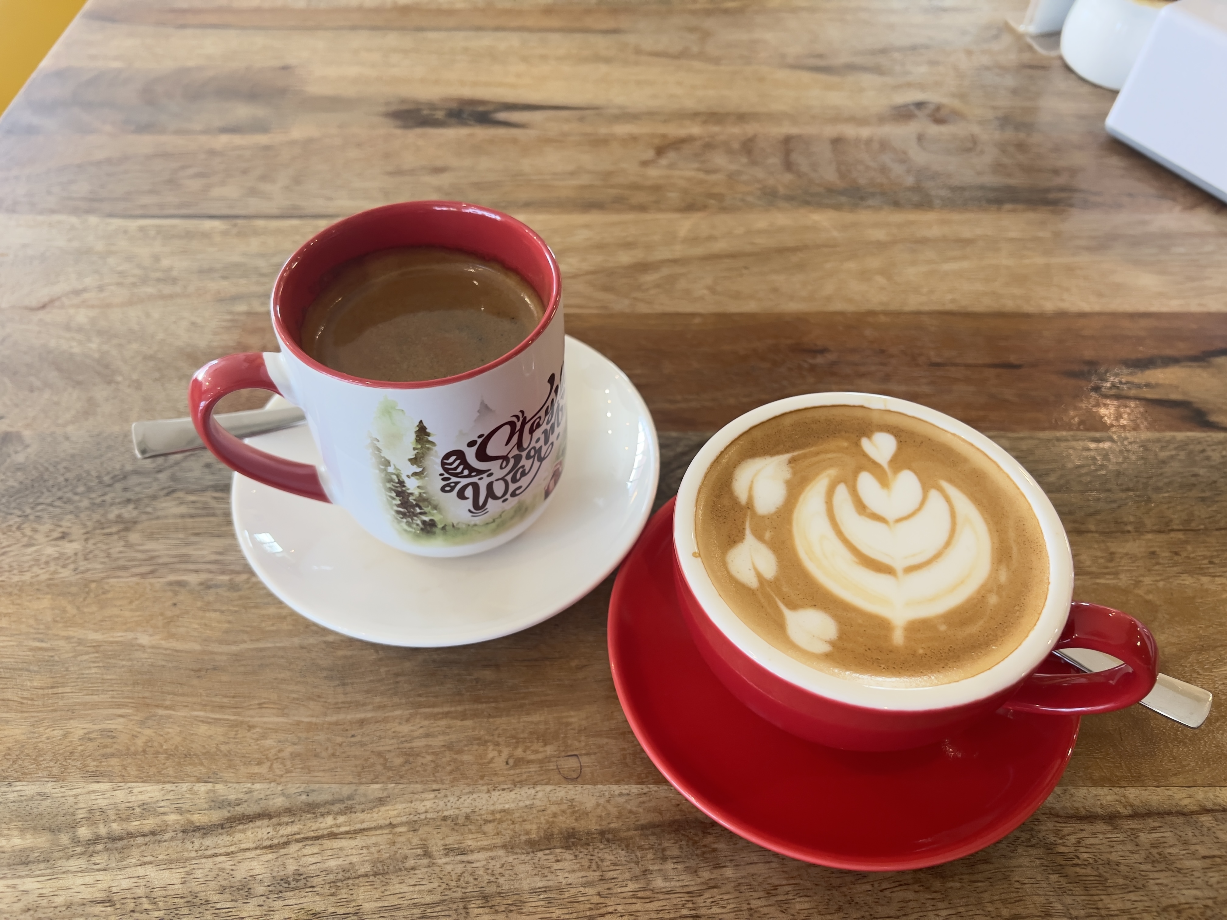 Two mugs with coffee sit on saucers on a wooden table. The mug on the left is white with a red rim and handle and has a print of a forest scene. The one on the right is red with a white saucer and has a detailed latte art design of a flower and hearts. 