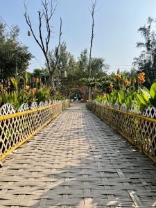 A narrow bamboo pathway leads through a lush garden, bordered by vibrant green plants and colorful flowers.