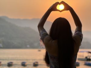 A silhouette of a person with their back to the camera, standing in front of a body of water with mountains in the distance. The person is holding their hands in a heart shape, with the setting sun visible through the heart.