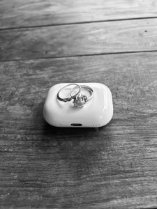 Two rings placed on top of a white wireless earbud case, set on a wooden surface in black and white.

