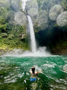 In a jungle gorge, a person swims in emerald water toward a tall waterfall that plunges into a misty pool, cliffs draped in lush green. Sunlit lens droplets halo the scene.