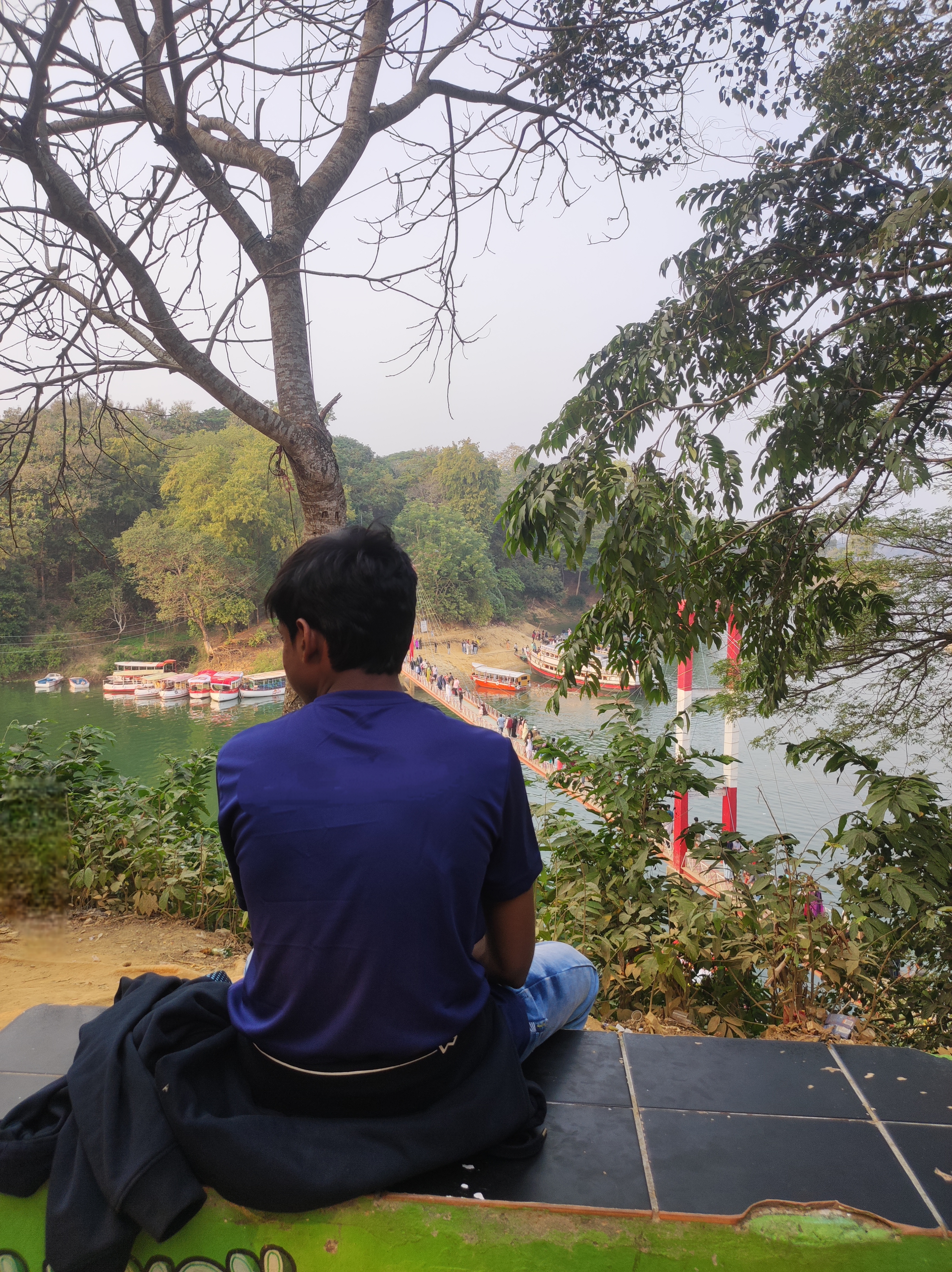 A person sits on a bench overlooking a river, facing away from the camera. The scene features a tree with bare branches and green foliage surrounding the area. In the background, a dock is visible, with several boats moored and a group of people walking along a bridge.