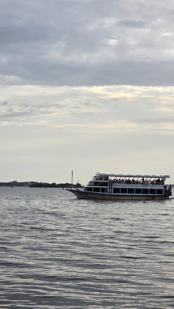 A large, multi-deck boat sails on a body of water under a cloudy sky. Captured from Kochi, Kerala.