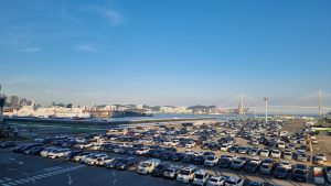 A panoramic view of Busan’s ferry terminal with rows of parked cars in the foreground, large ferries docked by the water, city buildings and a cable-stayed bridge visible across the bay under a clear blue sky. Busan International Passenger Terminal
