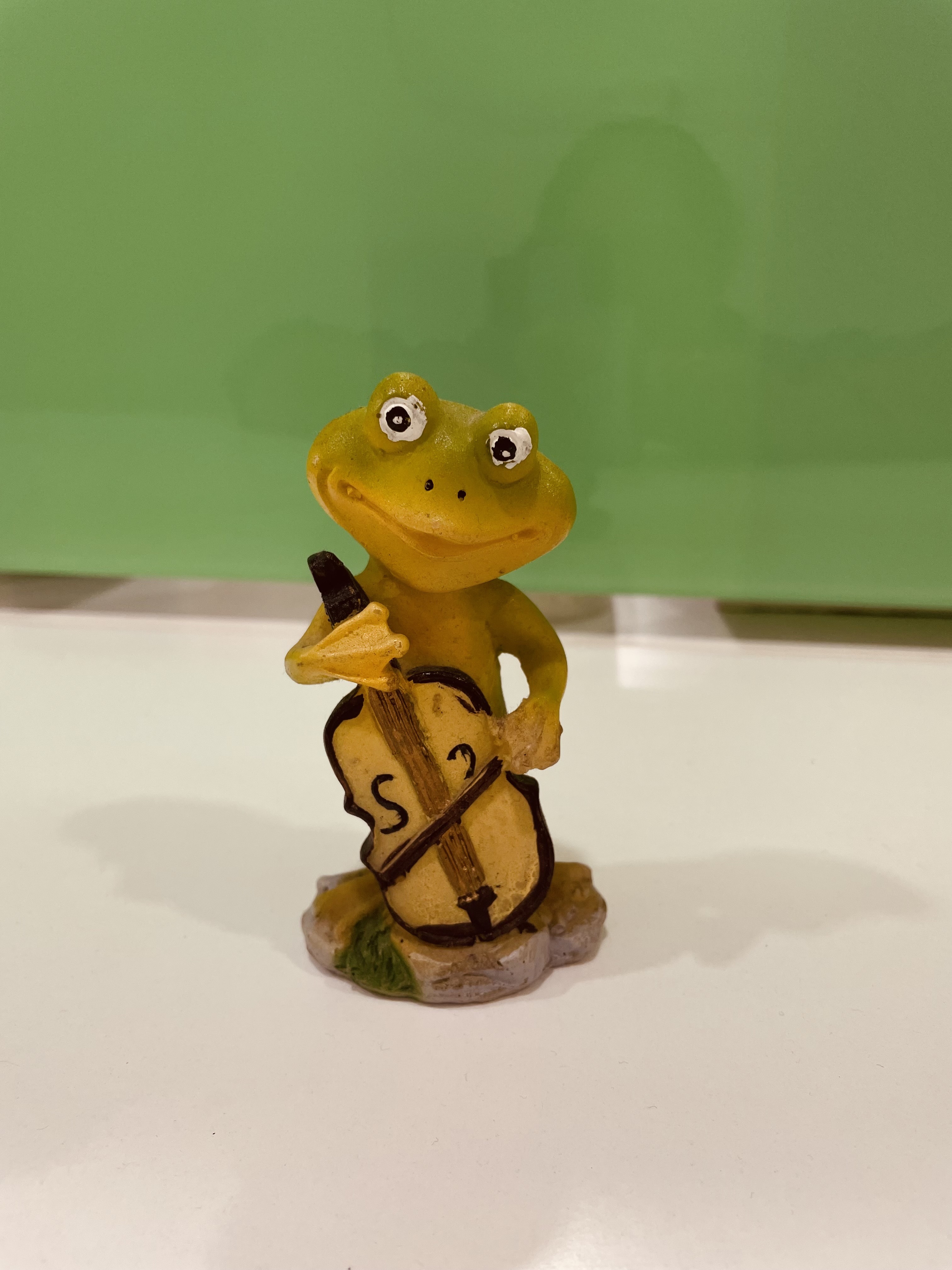 A whimsical yellow frog figurine is playing a cello, holding the instrument with one hand while the other hand holds a bow.