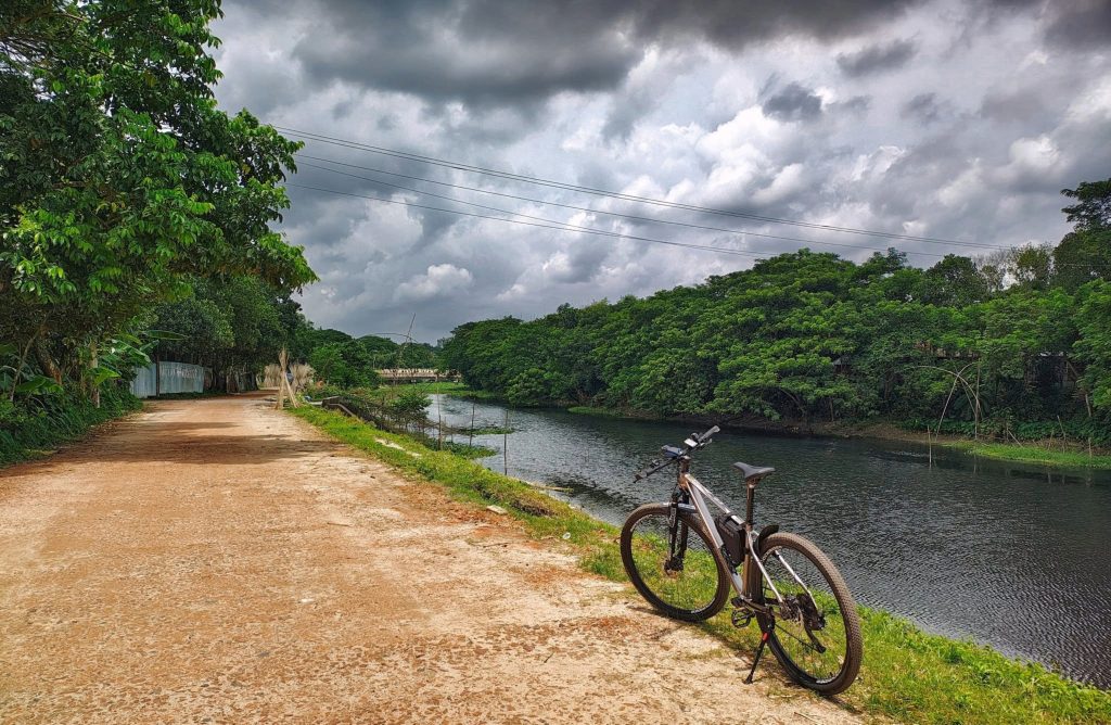 A scenic view of a dirt path alongside a river, with lush green trees on both sides. A bicycle is parked on the right side of the image, facing towards the water.