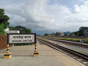 A railway station sign displaying "Akhaura Junction" in both Bengali and English stands on a platform. The scene shows railway tracks stretching into the distance, bordered by grassy areas and trees. Akhaura Junction, Bangladesh.