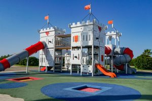A colorful playground featuring a castle-themed structure with two tower-like sections, connected by a red slide and a gray spiral slide. The playground has orange flags atop the towers and includes climbing elements, swings, and different textured surfaces in various colors 
