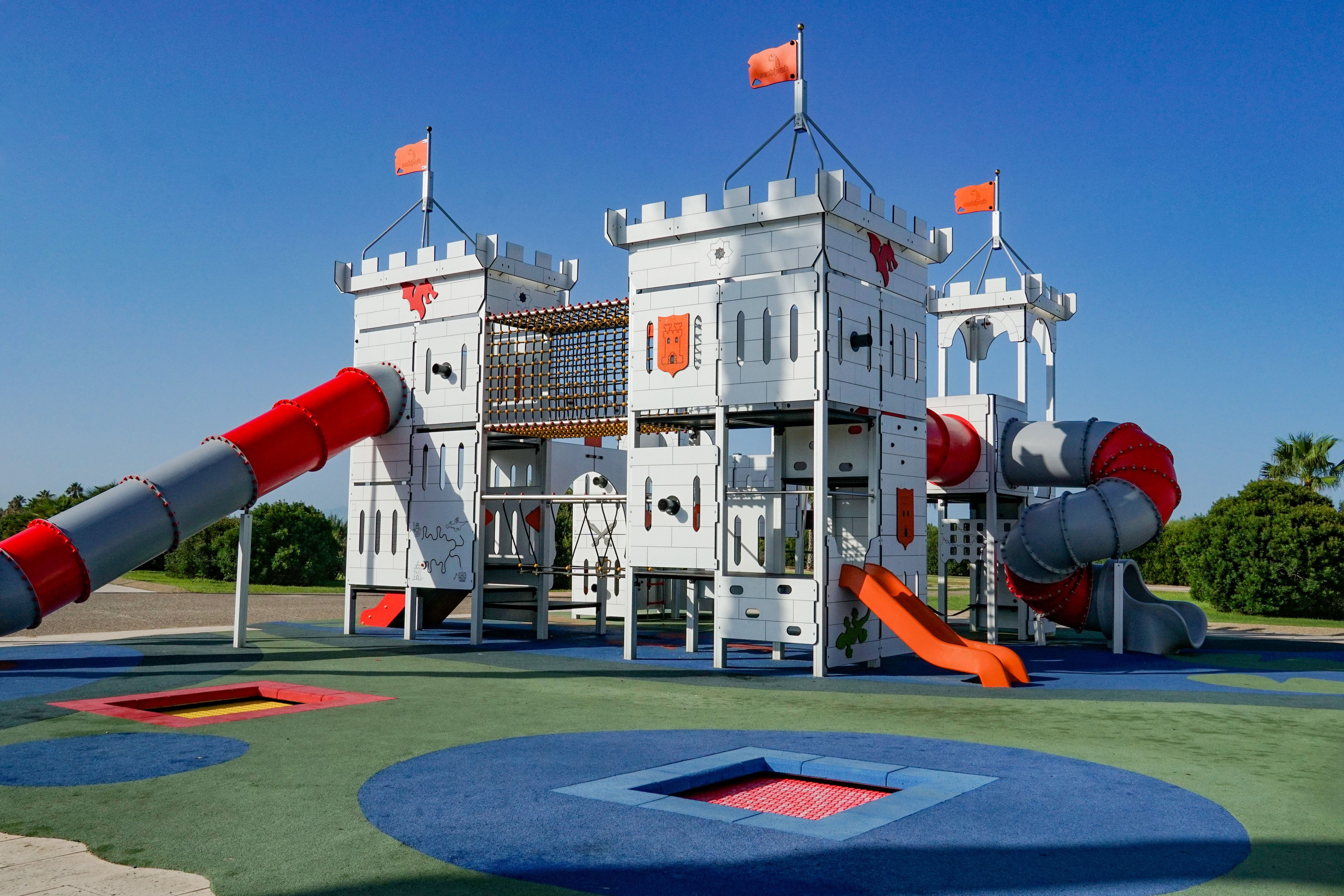 A colorful playground featuring a castle-themed structure with two tower-like sections, connected by a red slide and a gray spiral slide. The playground has orange flags atop the towers and includes climbing elements, swings, and different textured surfaces in various colors