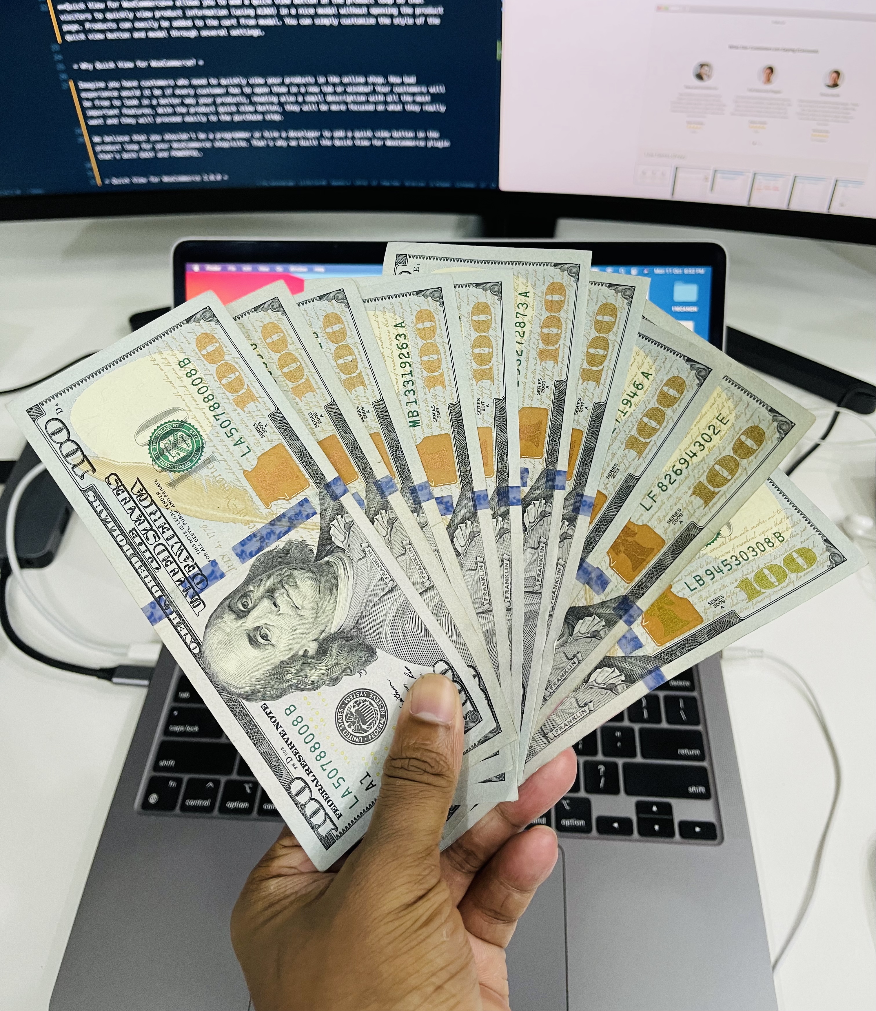 A person holding a fan of one hundred dollar bills over a laptop.