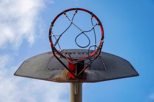 A close-up view of a street basketball hoop seen from below, with a weathered metal backboard and a partially rusted rim.