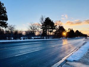 A snow-covered road lined with trees, viewed during sunset.
