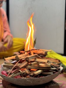 A small ritual fire burns in a shallow clay bowl, which is resting on two bricks. A person wearing a yellow and orange A close-up view of a fire burning atop a stacked pile of wooden sticks arranged in a circular stone bowl. The flames are vividly orange and yellow, creating a warm glow. In the background, a person dressed in traditional attire is sitting, partially out of focus, with yellow fabric visible, suggesting a cultural or ceremonial setting. The ground is adorned with colorful fabric, adding to the ambiance.