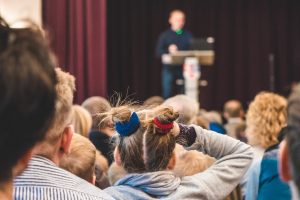 Audience listening to a speaker on stage, with the focus on a child in the crowd wearing two hair buns tied with red and blue scrunchies.