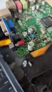  A close-up image of a green printed circuit board featuring various electronic components, including capacitors, resistors, and connectors. Some of the components are labeled with values. In the background, there are blurred out tools and screws, suggesting a workspace environment focused on electronics repair or assembly.

