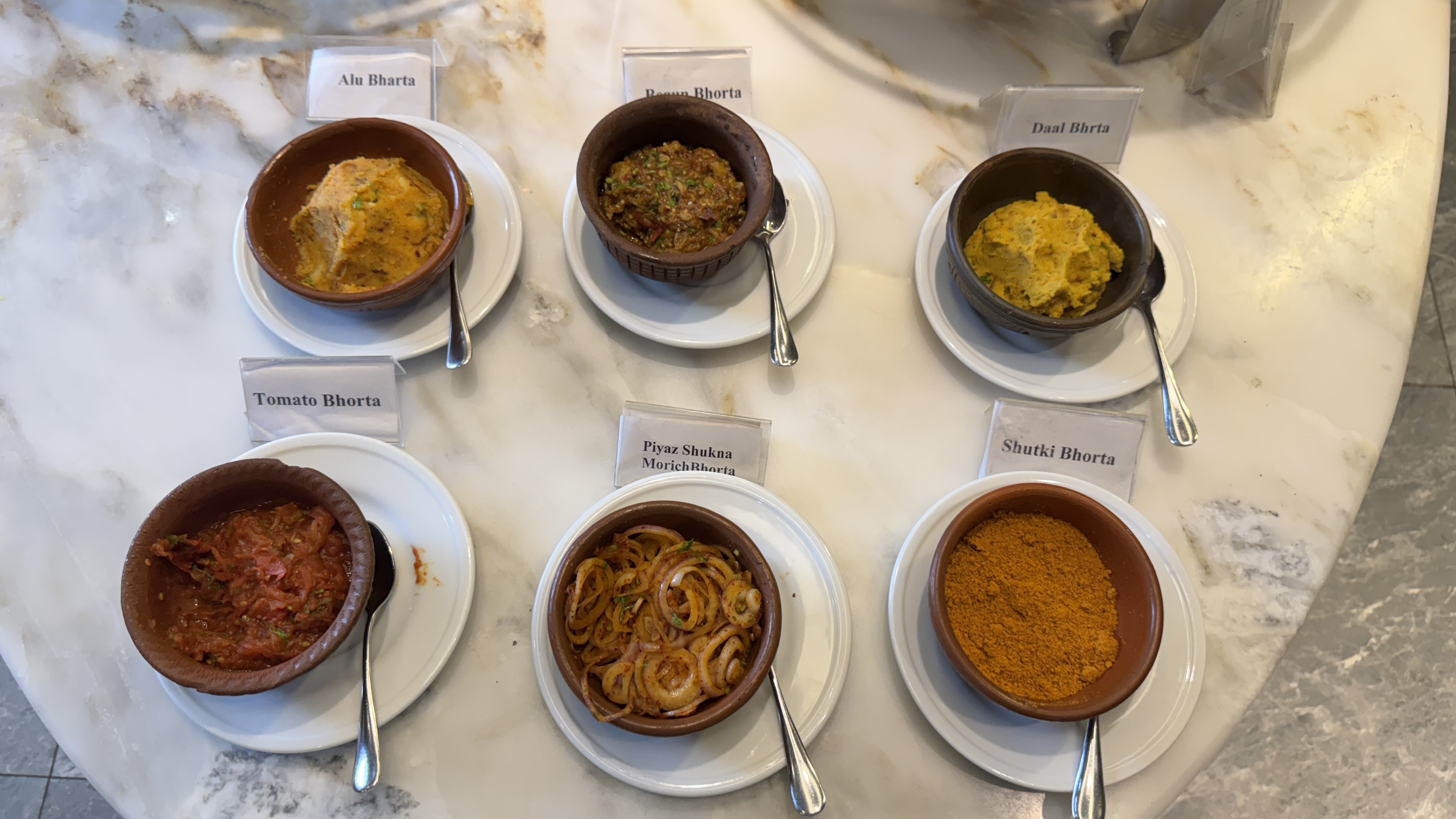 Six small brown bowls sit on a white marble surface, each filled with a different Bangladeshi vorta—mashed potato, lentil, tomato, eggplant, spicy onion, and dried fish powder.