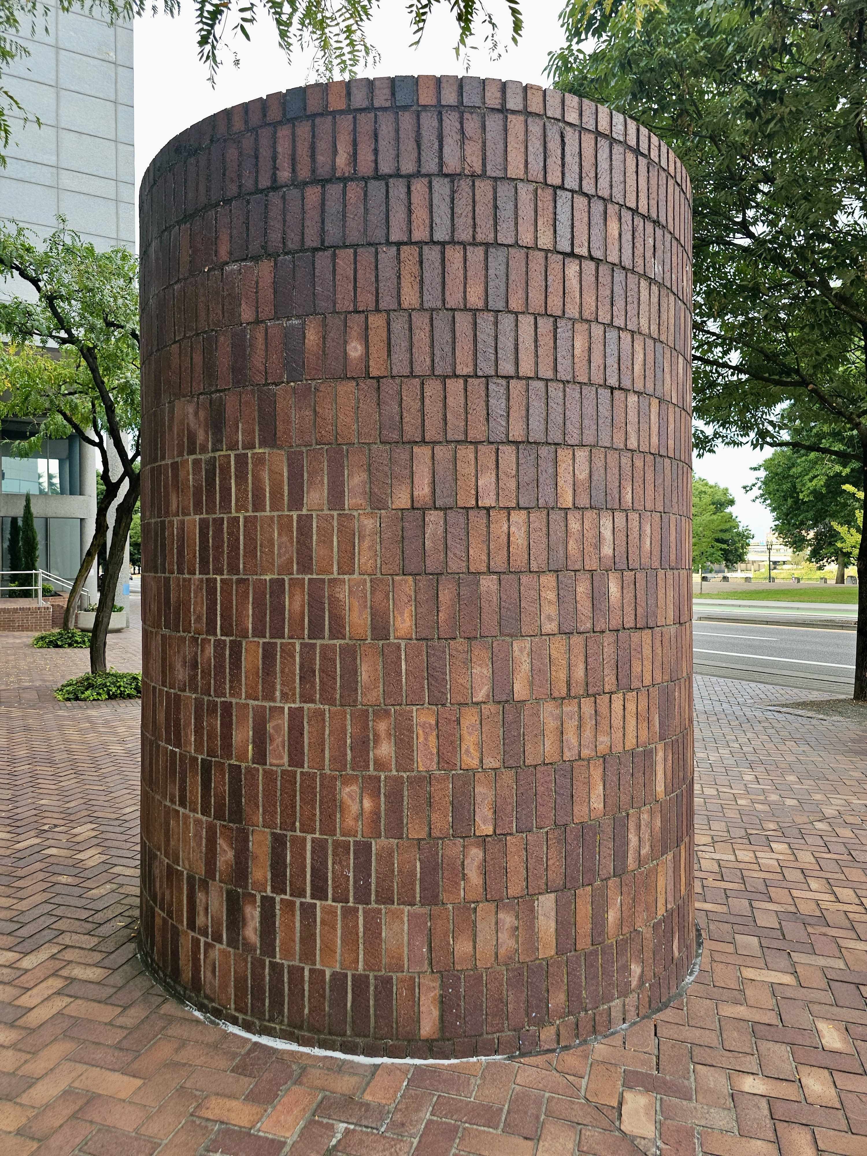 A cylindrical structure made of reddish-brown bricks with a textured surface, standing outdoors amidst some greenery and paving. Captured from Portland