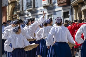 Group of women in traditional Basque fisherwoman attire take part in the Basque festival parade in Irun, Spain. They wear white blouses, dark blue skirts, and blue-and-white checkered headscarves, carrying trays for fish. One of the women waves to the crowd.