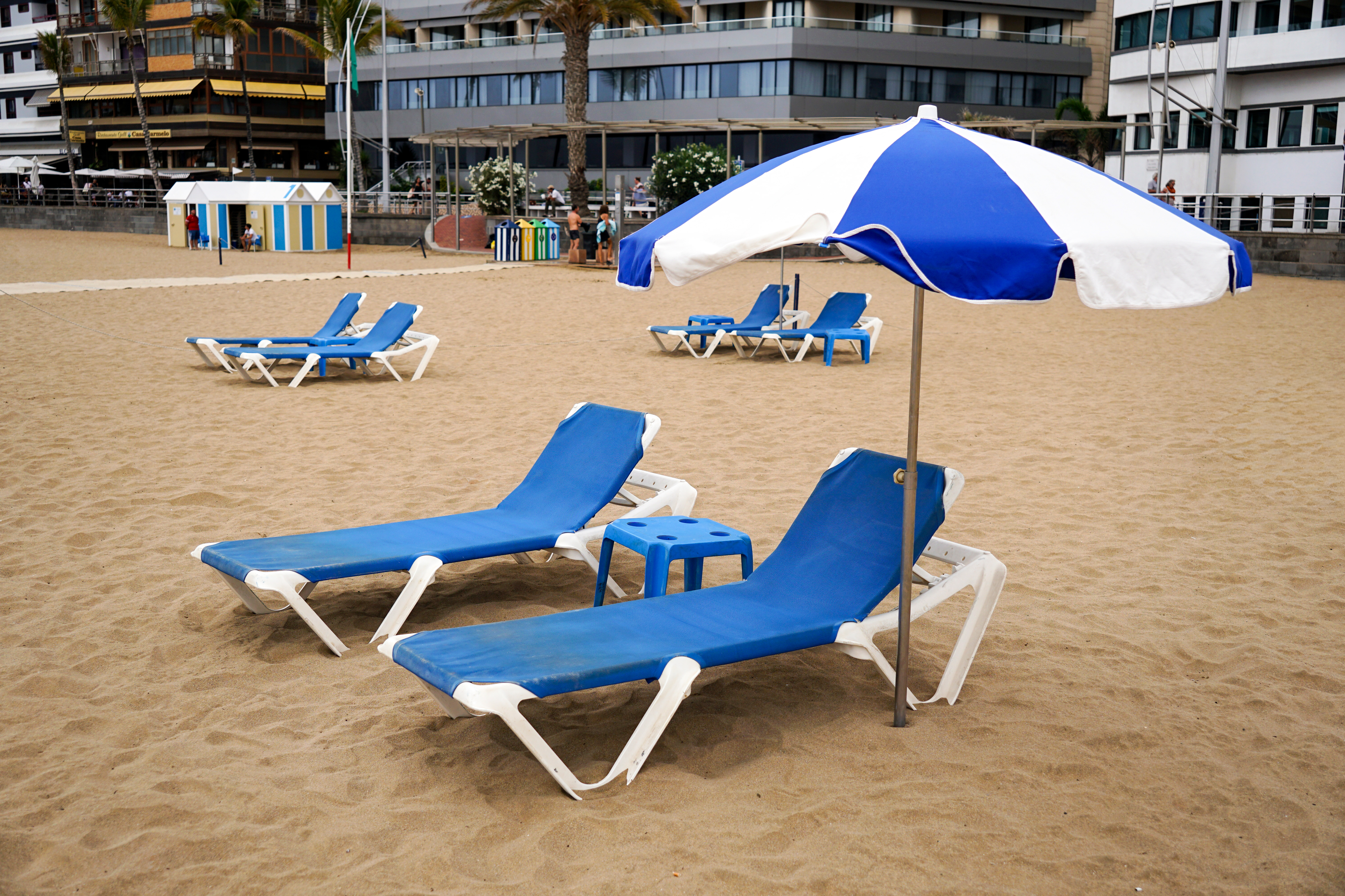 A beach scene featuring several blue lounge chairs set on sandy ground. A blue and white striped umbrella shades one of the chairs, and a small blue side table is nearby. In the background, there are more lounge chairs, a kiosk with colorful walls, and buildings with palm trees.