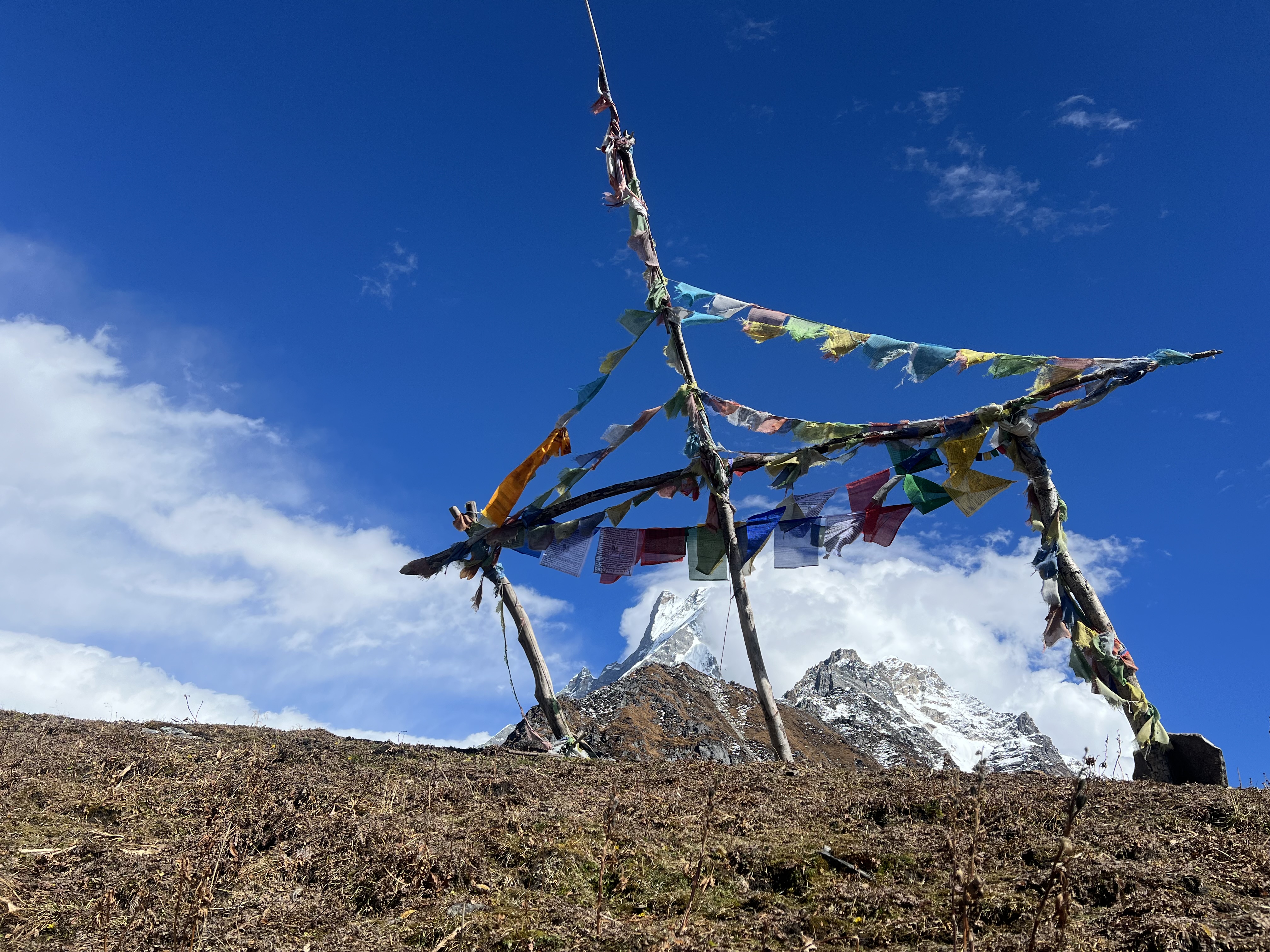 A traditional prayer flag structure on a rocky, brown hill, with a snow-capped mountain peak and a clear blue sky in the background.