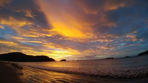 Sunset on the beach with the sky painted in intense shades of orange, gold, and blue, reflected on the wet sand and calm sea, with mountains and boats on the horizon.
