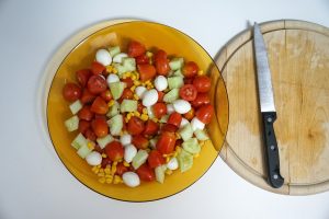 A yellow bowl filled with a colorful salad featuring cherry tomatoes, small mozzarella balls, diced cucumber, and sweet corn. The bowl is placed on a white surface next to a wooden cutting board with a knife resting on it. 