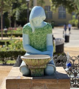 Sunlit water fountain. It consists of a figure and a container. The figure represents a person sitting on their lap, looking down. She has long hair, a short-sleeved green sweater, and a skirt. A hand touches the container. In the background, blurred, there is a building, vegetation, and some people walking.