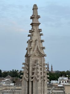 Close-up of a tall, ornate stone spire against low-rise buildings and an overcast sky.