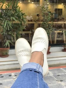 A person's feet are crossed, wearing blue jeans and white perforated loafers, with potted plants and a cafe in the blurry background.