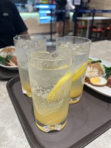 Three tall glasses of a fizzy, light-colored drink with lemon slices and ice are on a brown tray. 