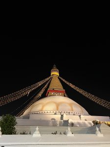 A Buddhist stupa is silhouetted against a colorful sunset. The stupa is decorated with prayer flags and a golden spire