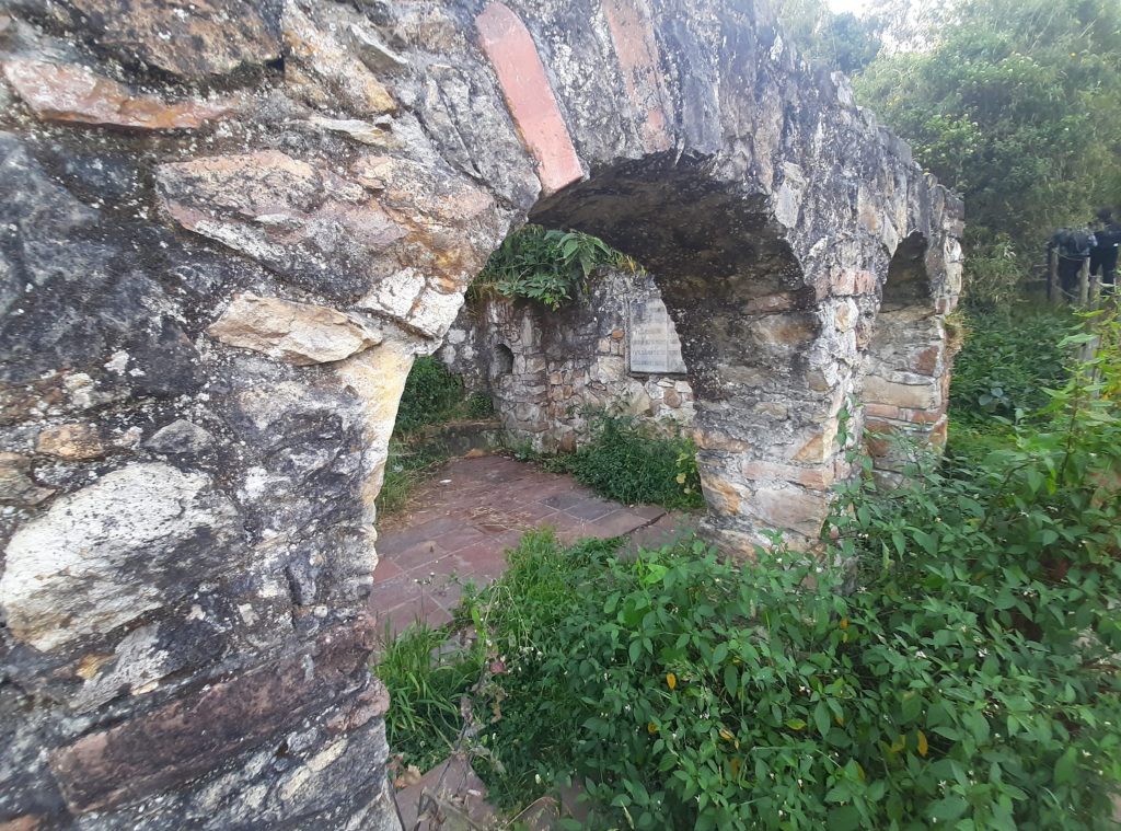 A stone structure with multiple arches partially covered in greenery, revealing a brick floor and surrounding wild plants. The walls show signs of weathering and age, indicating the ruins of an old building or facility. A faintly visible plaque rests against one wall, adding to the historical ambiance of the site.