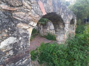 A stone structure with multiple arches partially covered in greenery, revealing a brick floor and surrounding wild plants. The walls show signs of weathering and age, indicating the ruins of an old building or facility. A faintly visible plaque rests against one wall, adding to the historical ambiance of the site.