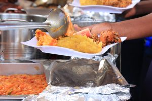 A close-up of a hand holding a disposable plate being served with food at a buffet. The plate contains matoke (mashed plantain), fried rice, stewed chicken, vegetables, and a slice of watermelon, with more food being ladled onto it. Shiny foil trays and saucepans are visible in the background.