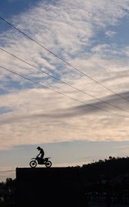 Silhouette of a man on a motocross bike atop a platform against a cloudy sunset sky.