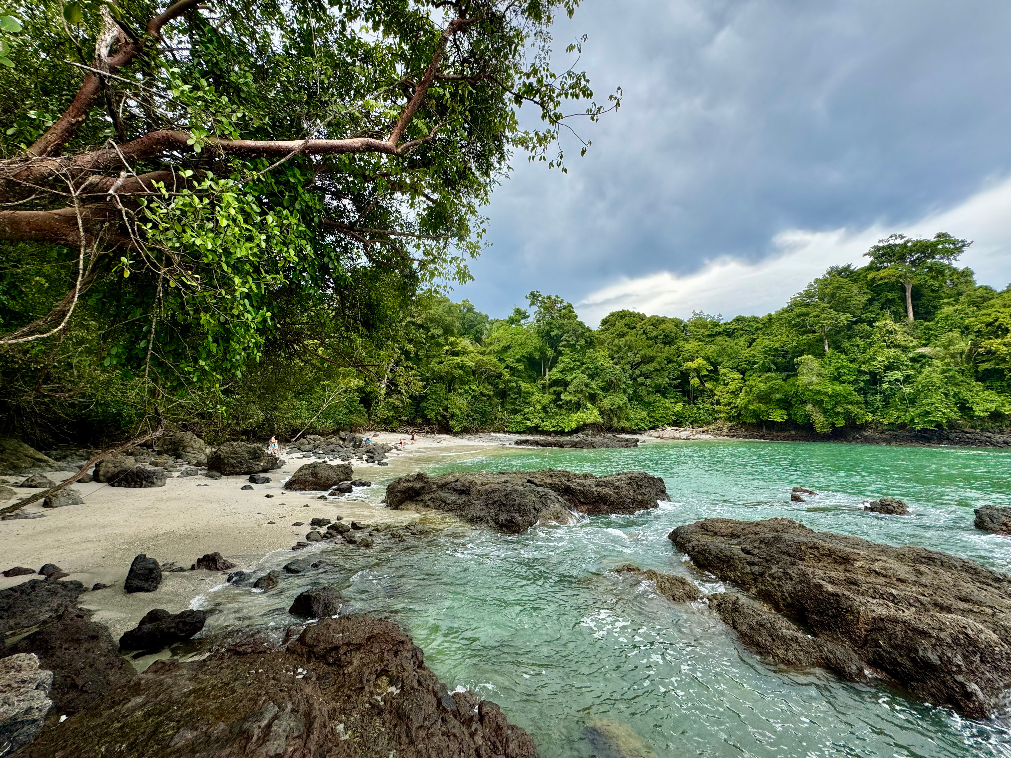 
Turquoise waters lap dark volcanic rocks and a small sandy beach in a jungle-fringed cove, framed by overhanging branches beneath a cloud-heavy sky.