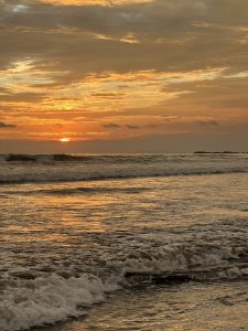 A serene beach scene at sunset, featuring golden and orange hues in the sky as the sun dips below the horizon. Gentle waves roll onto the shore, reflecting the warm colors of the sky, while the outlined silhouette of distant rocks is visible in the water. Playa Jacó, Costa Rica