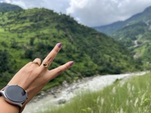 A hand with a smartwatch and two rings makes a peace sign in front of a river and green mountains