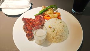 A delicious-looking meal is presented on a white circular plate. The plate contains a seared piece of meat, a serving of white rice, and a side of mixed vegetables including carrots, cauliflower, and green beans.