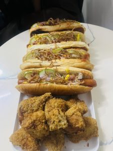 A large serving plate with several homemade hot dogs lined up next to a pile of fried chicken pieces. The hot dogs are topped with onions, pickles, and mustard. 