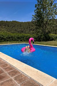 Swimming pool with terrazzo and stone edge on the left, a person entering a pink flamingo float with water splashing around, and green vegetation and clear blue sky in the background.
